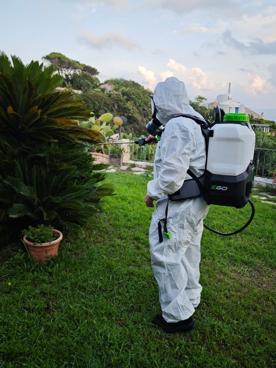 Fulvio Perrone in tuta bianca con cappuccio e maschera che nebulizza con una pompa a spalla la vegetazione di un giardino affacciato sul mare, a Gaeta